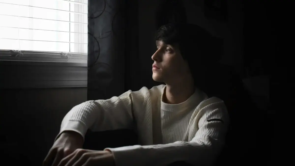 man wearing white crew-neck sweater and black knit cap beside black curtain