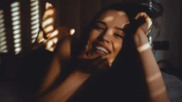 A smiling woman lies on a bed with striped shadows