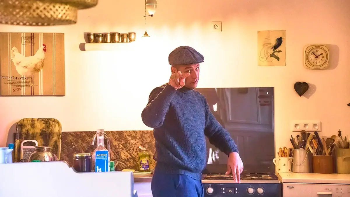 Man in flat cap talking on phone in kitchen.