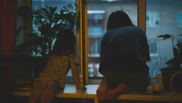 A woman sitting by a kitchen window in soft morning light, hands folded, thoughtful