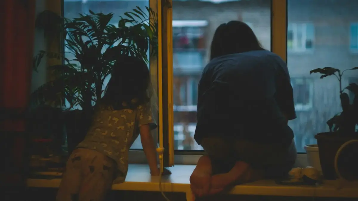 A woman sitting by a kitchen window in soft morning light, hands folded, thoughtful