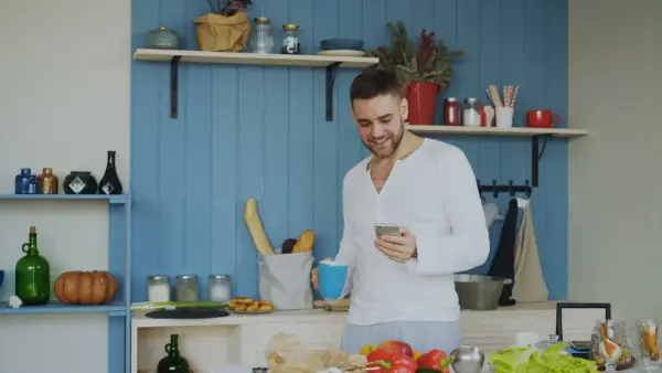 Man smiling while holding coffee and phone in kitchen.