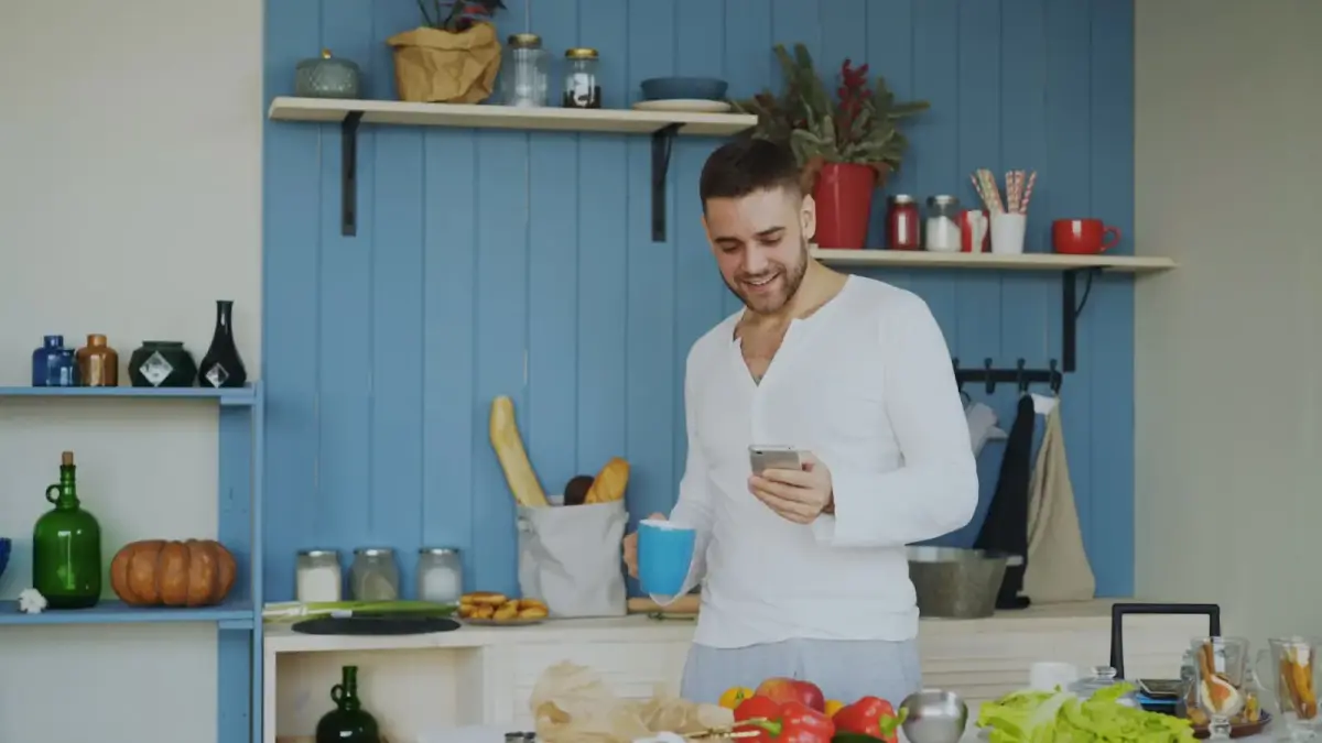 Man smiling while holding coffee and phone in kitchen.