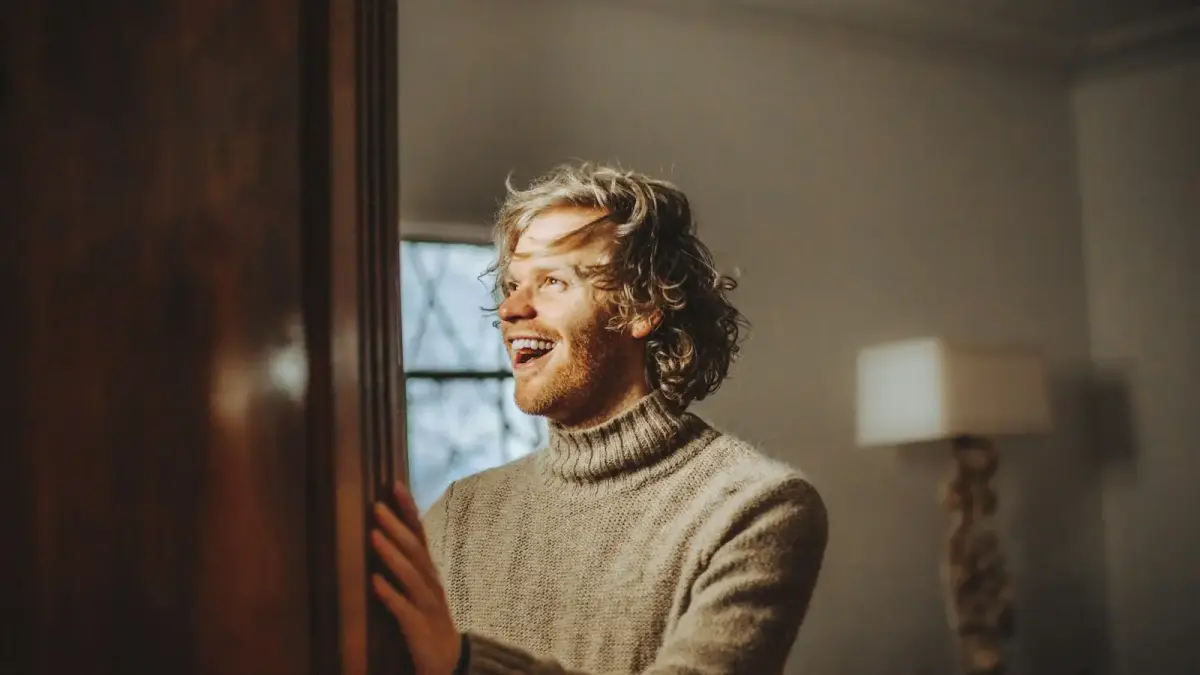man wearing brown sweater standing and smiling near wall