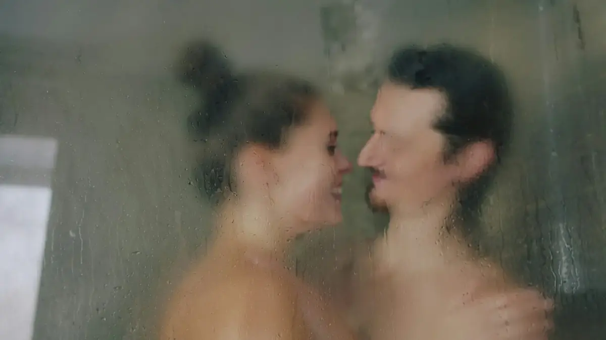 A woman and a man facing each other behind a wet glass shower door