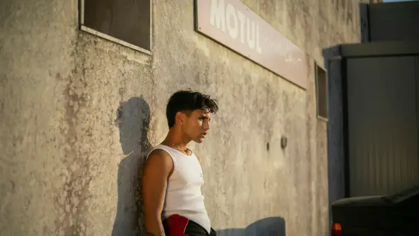 Man in tank top leans against a weathered wall.