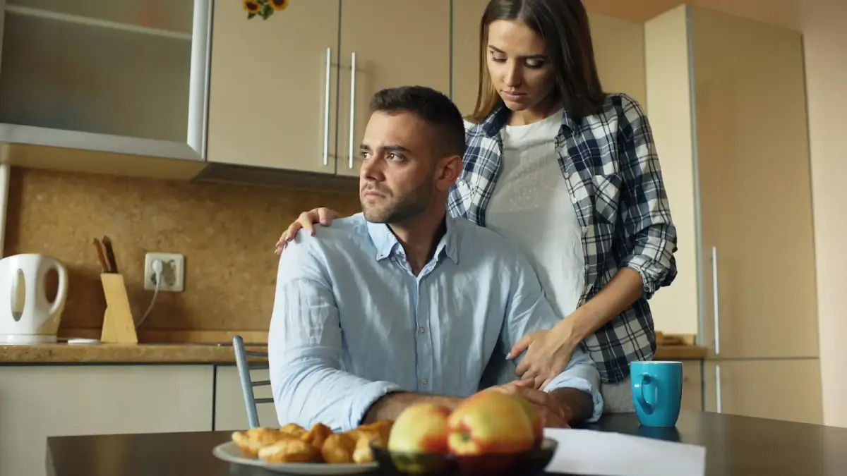 Woman comforts man at kitchen table with food.