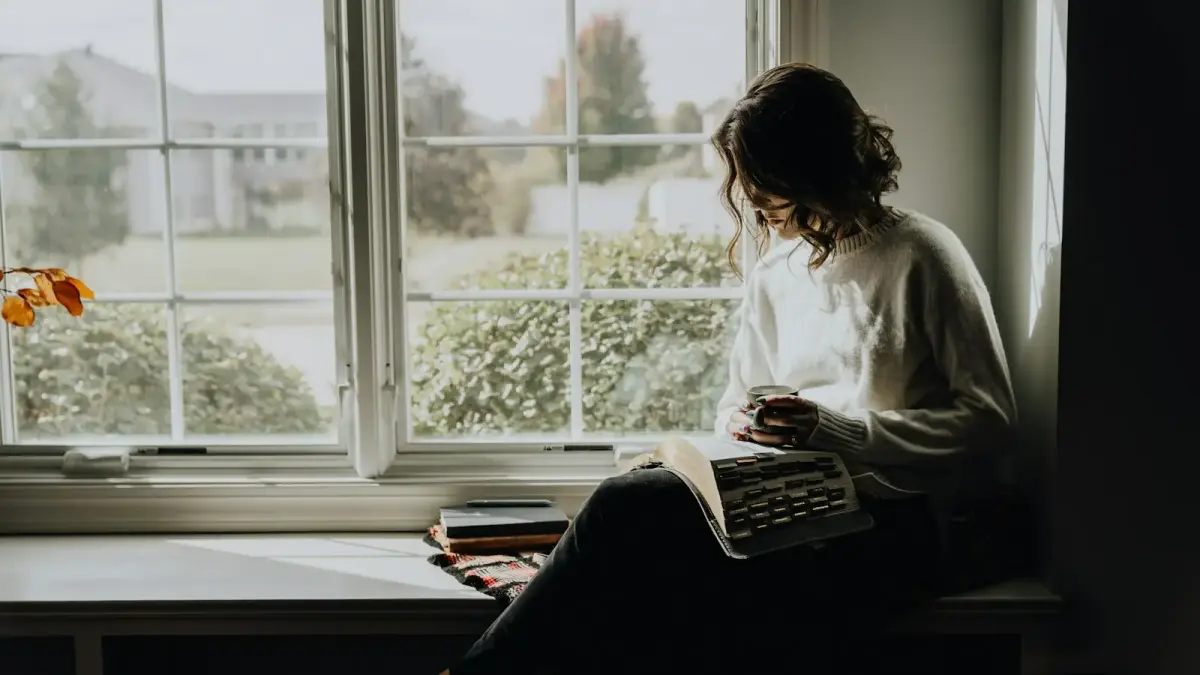 A woman in a white sweater holding a mug while reading a book